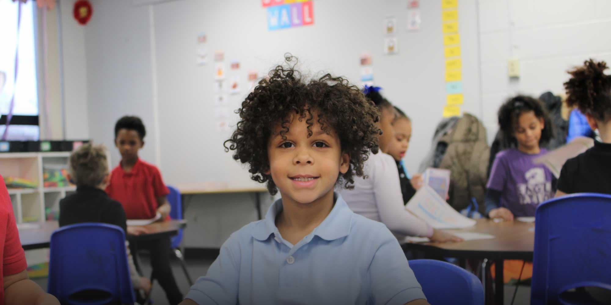 Smiling student with curly hair sitting in class with classmates in the background.