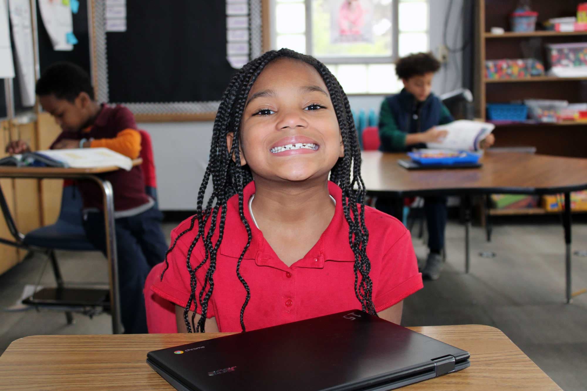 Smiling student in a red shirt at a desk with a Chromebook.