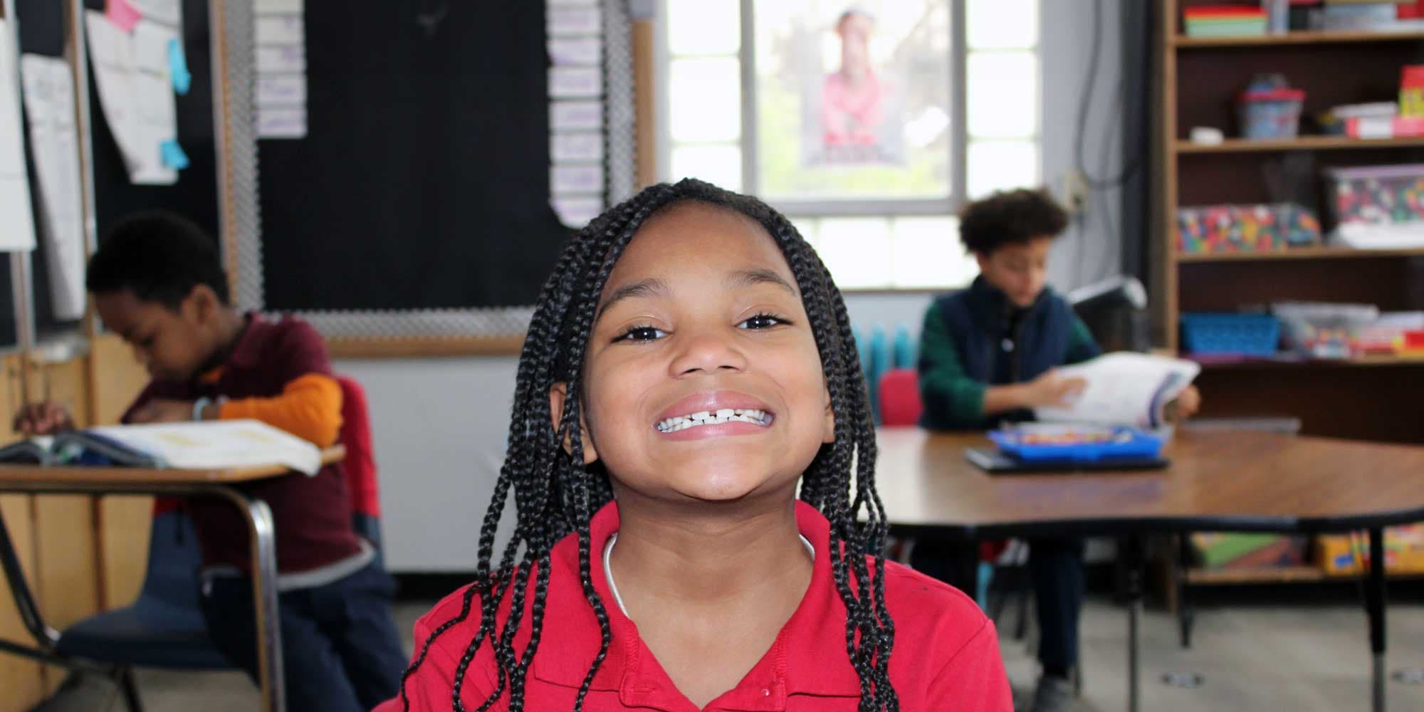 Smiling student in a classroom while classmates read at their desks.