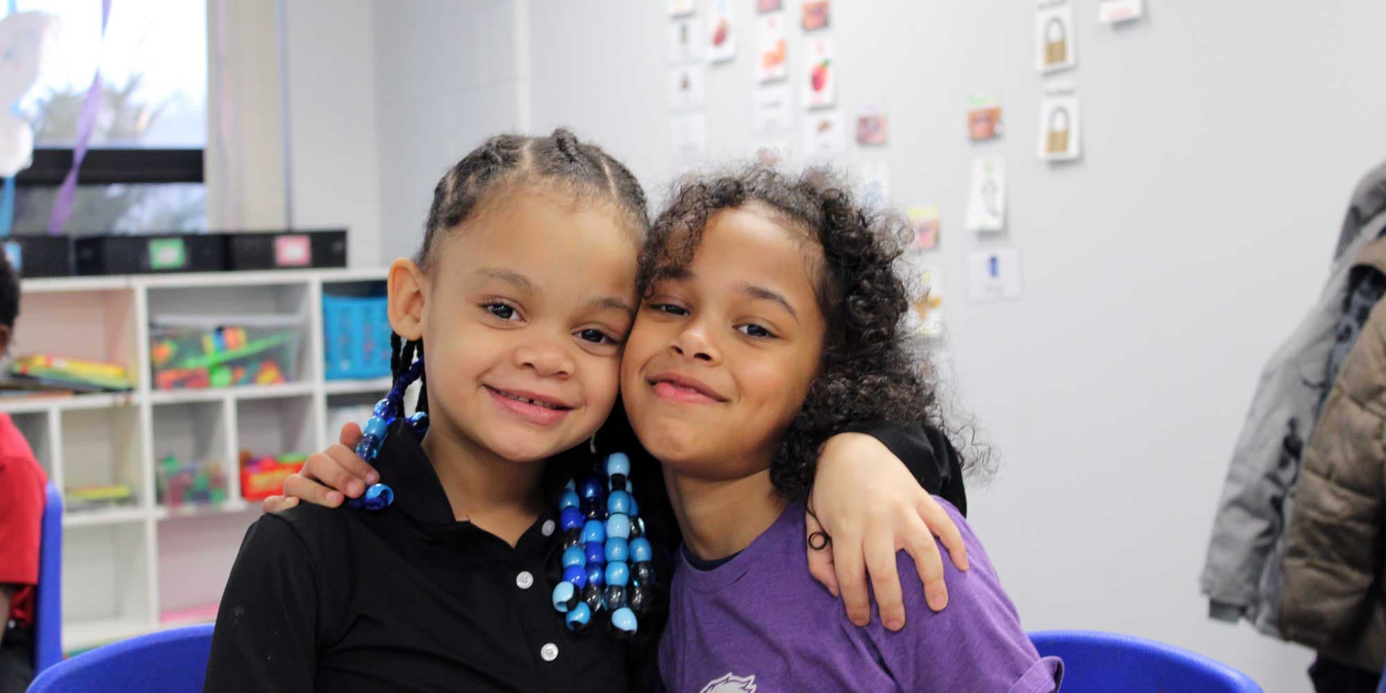 Two smiling students with their arms around each other in a classroom.